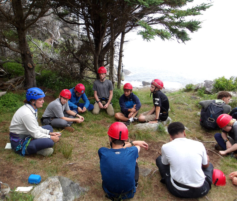 A group of people wearing helmets are sitting in a circle on a grassy area near trees and a body of water. They appear to be engaged in a discussion or activity, possibly related to outdoor adventure or team building. The helmets suggest a focus on safety, and the setting implies an outdoor or recreational context.